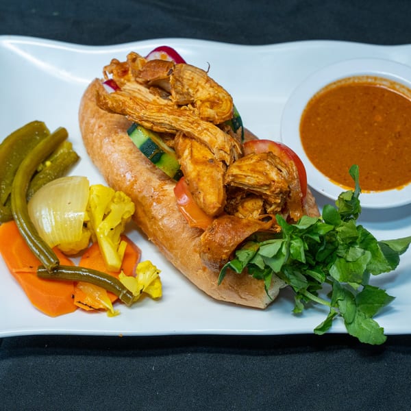 A Salvadoran chicken sandwich with vegetables and a side of red sauce on a white plate.