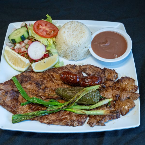 Grilled steak with sausages, green onions, rice, beans, and a fresh salad with lemon wedges.