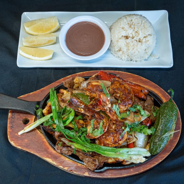 Sizzling fajitas with shrimp, beef, peppers, and onions, served with rice, beans, and lemon wedges.