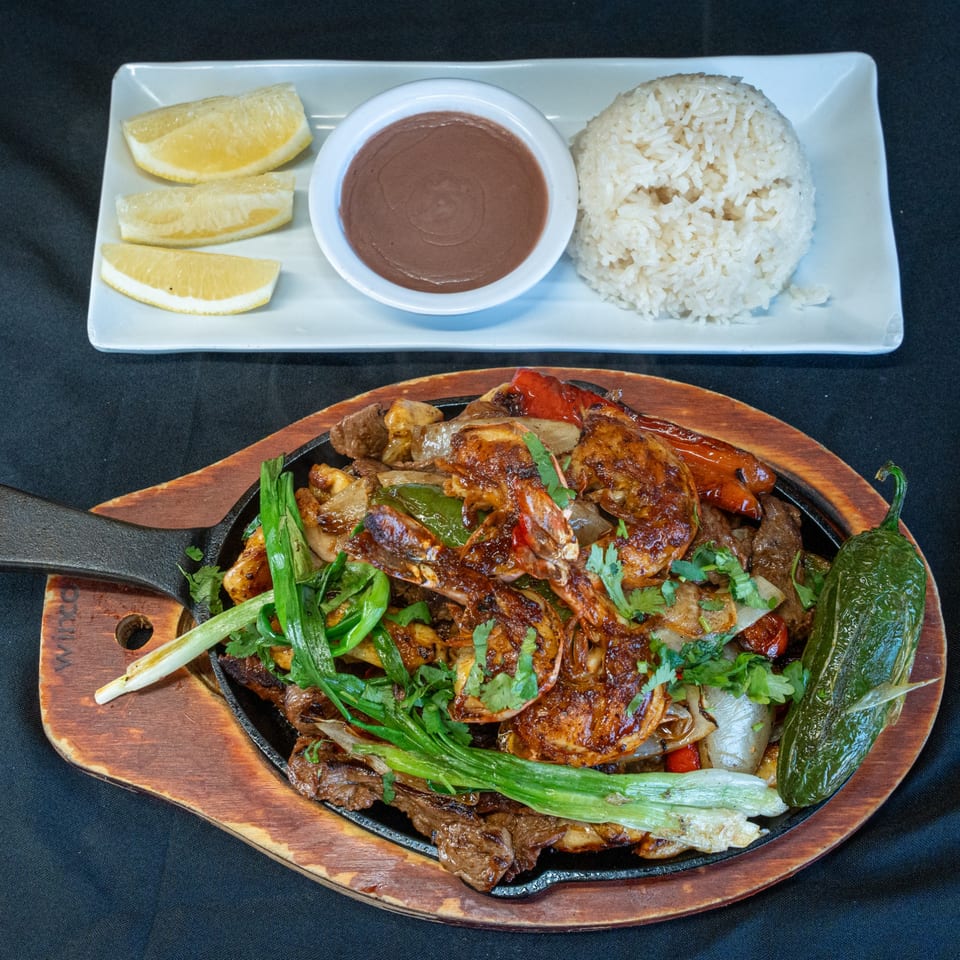Sizzling fajitas with shrimp, beef, peppers, and onions, served with rice, beans, and lemon wedges.